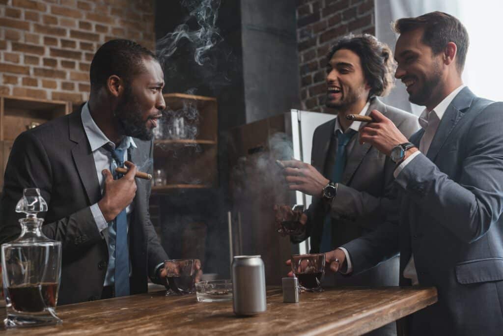 smiling multiethnic male friends in suits smoking cigars, drinking whiskey and talking