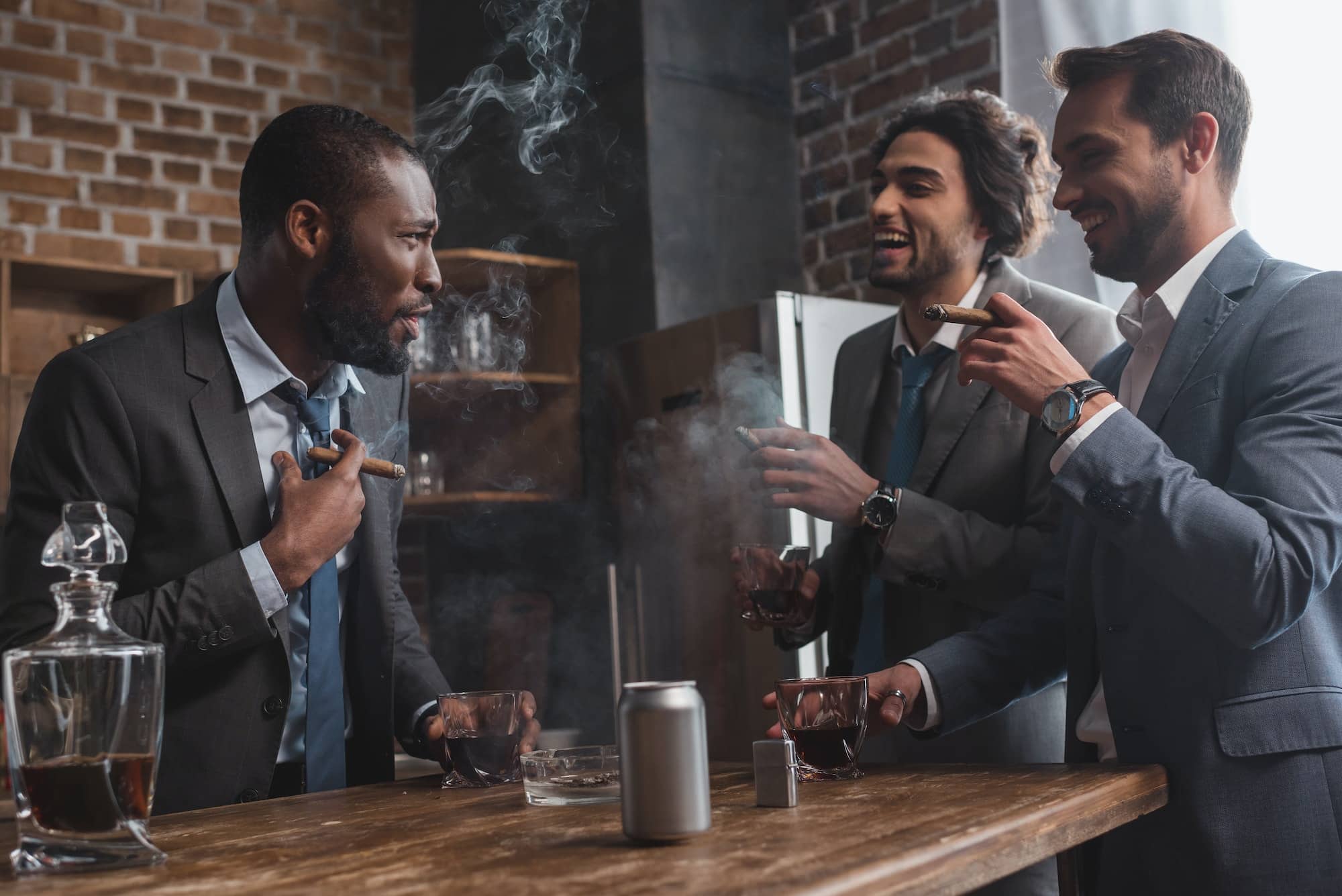 smiling multiethnic male friends in suits smoking cigars, drinking whiskey and talking