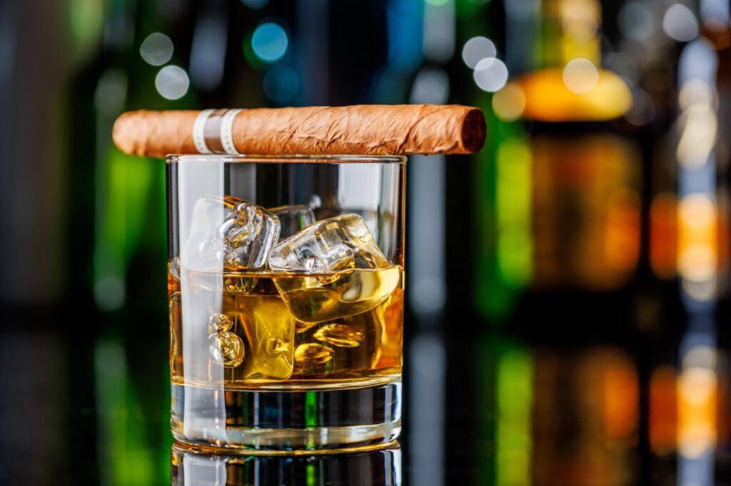 The Cigar Store Whiskey on the rocks beside a lit cigar on a bar table
